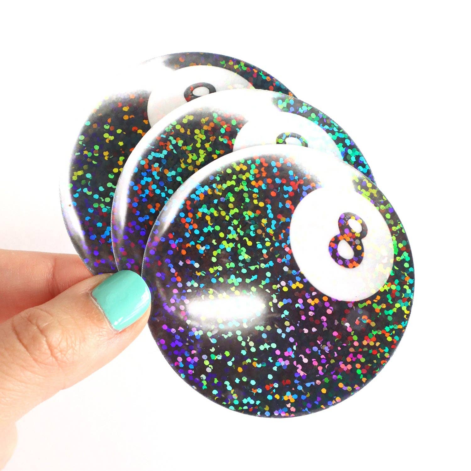 Three glitter 8 ball stickers held by a hand with turquoise nail polish on a white background.