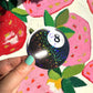 Hand holding a glittery eight-ball sticker in front of colorful strawberry illustrations.