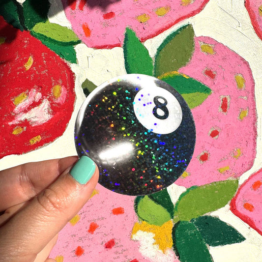 Hand holding a glittery eight-ball sticker in front of colorful strawberry illustrations.
