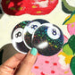 Three glittery '8' ball stickers held by a hand with green nail polish against a colorful background.