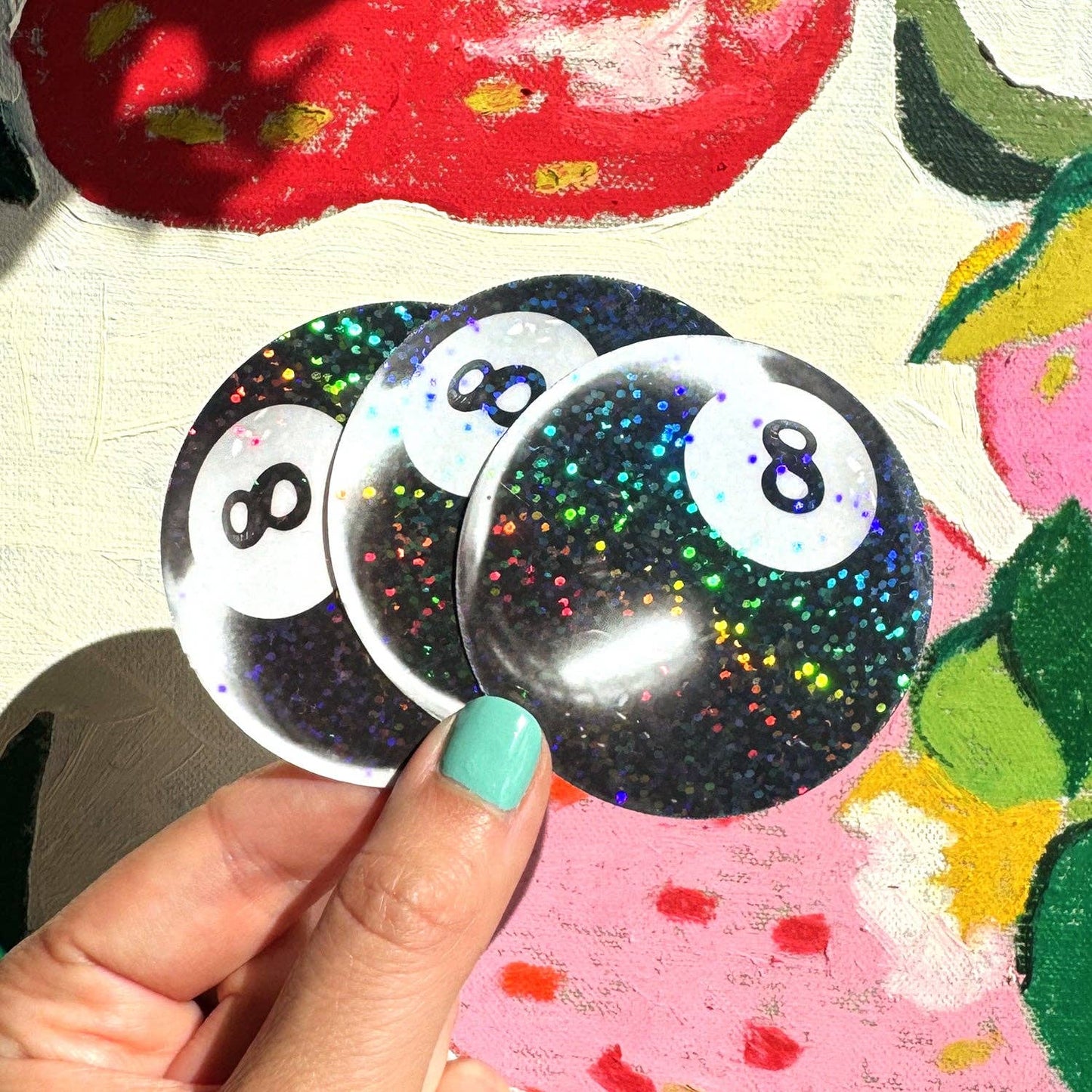 Three glittery '8' ball stickers held by a hand with green nail polish against a colorful background.