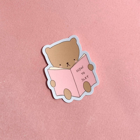 Sticker of a bear holding a book titled 'How to Slay' on a pink background