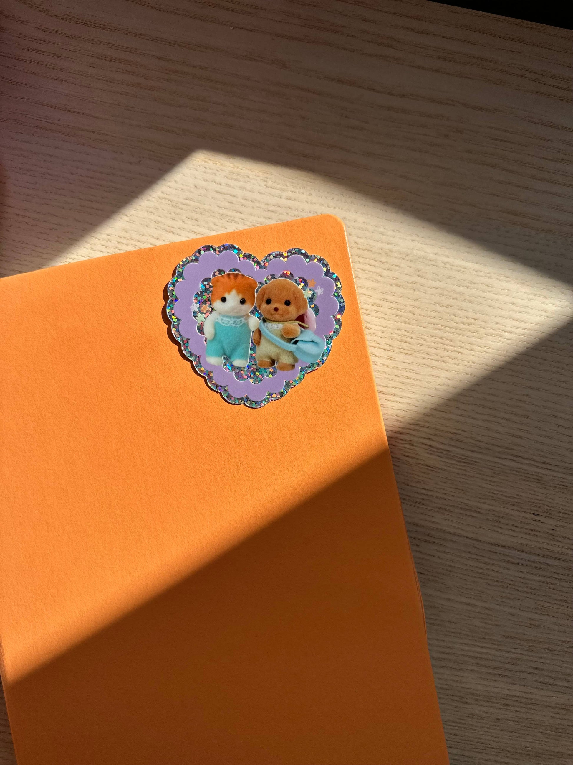 Orange background with a heart-shaped sticker featuring two sylvanian characters on a wooden surface.