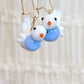 A pair of handmade earrings featuring small white and light blue bird charms with orange beaks and black eyes, suspended from gold-tone wire hooks against a soft beige background.