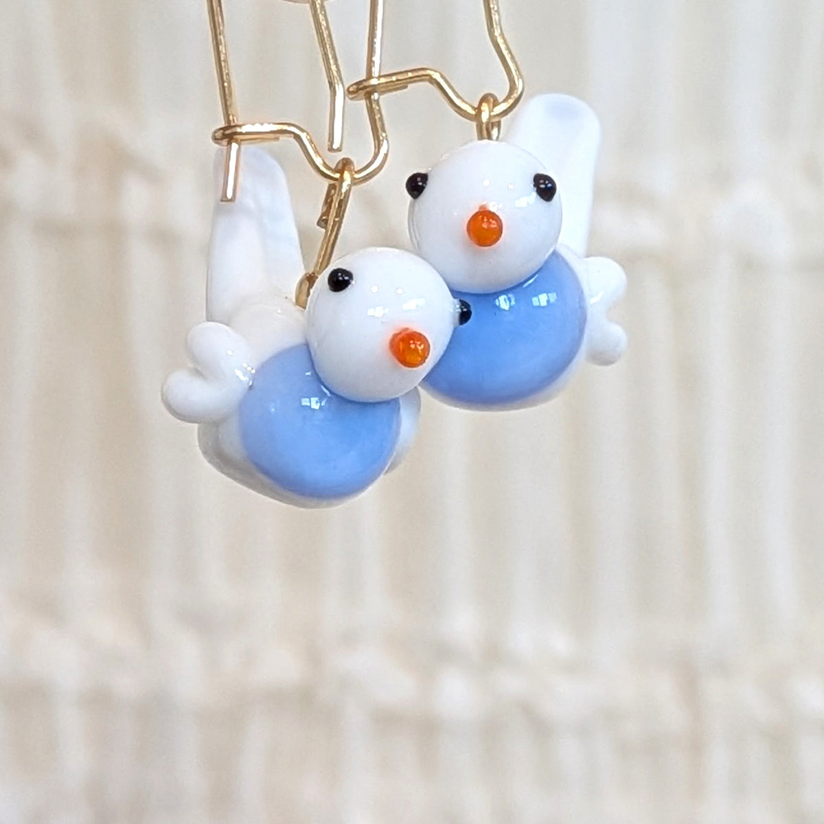 A pair of handmade earrings featuring small white and light blue bird charms with orange beaks and black eyes, suspended from gold-tone wire hooks against a soft beige background.