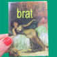 magnet of a classical painting and the word 'brat' in green text, held by a hand with red nail polish against a green background.