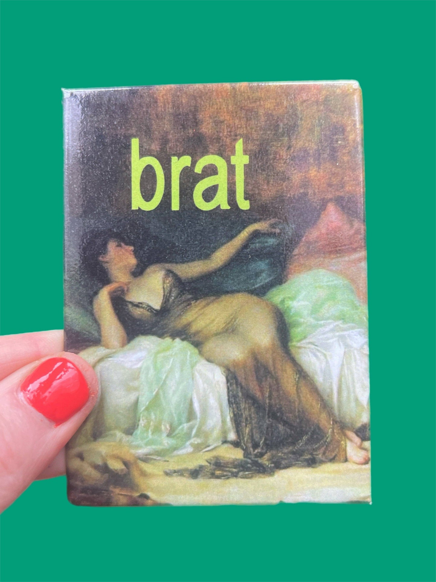 magnet of a classical painting and the word 'brat' in green text, held by a hand with red nail polish against a green background.