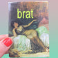 magnet of a classical painting and the word 'brat' in green text, held by a hand with red nail polish.