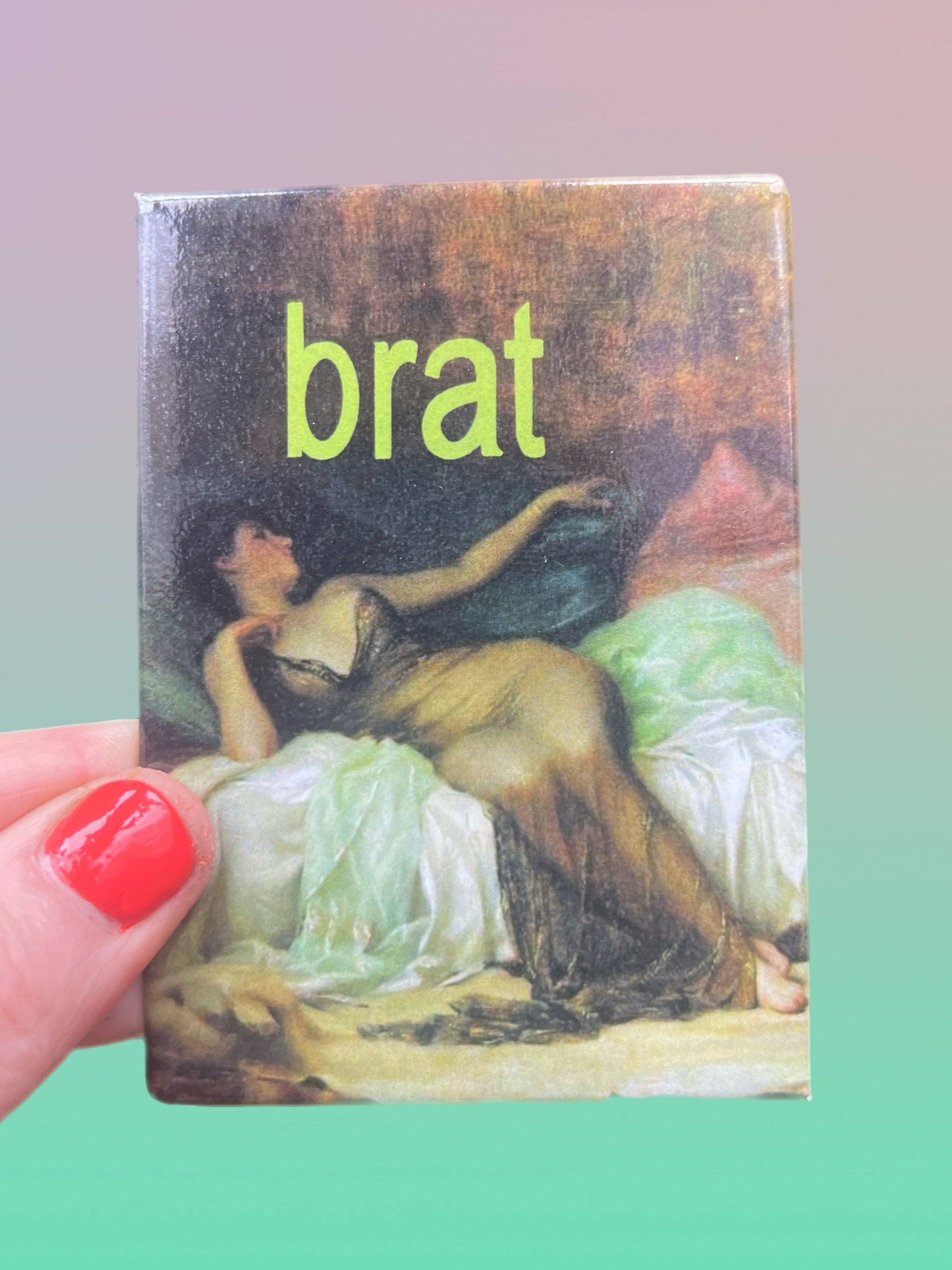 magnet of a classical painting and the word 'brat' in green text, held by a hand with red nail polish.