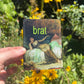 Hand holding a sticker with a classical painting and the word 'brat' in green text, surrounded by flowers.