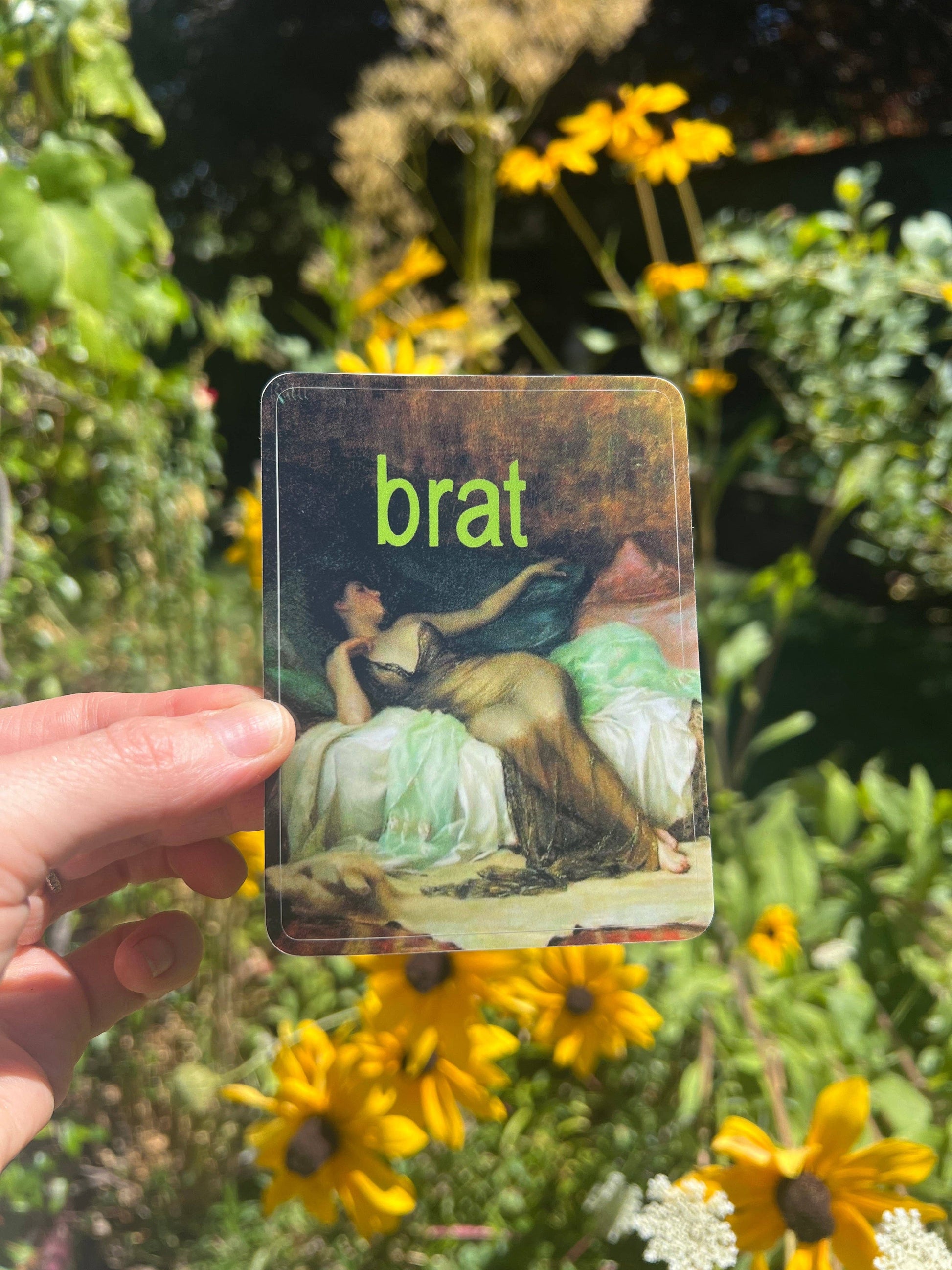 Hand holding a sticker with a classical painting and the word 'brat' in green text, surrounded by flowers.