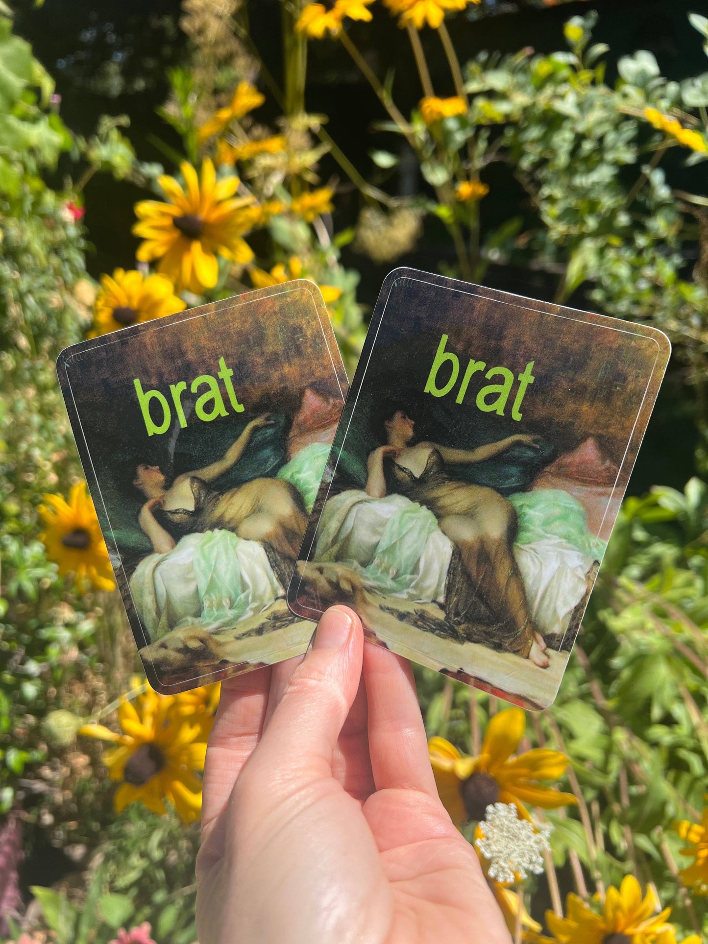 Two stickers  labeled 'brat' held in front of a natural background with flowers.