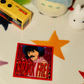 A square sticker  with the words 'BREAK FREE' in large red letters, placed on a white surface with star-shaped cutouts and small plush toys nearby.
