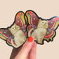 Butterfly-shaped sticker with a cat design held by a hand against a beige background