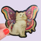 Sticker of a cat with butterfly wings held by a hand with red nail polish on a light purple background.
