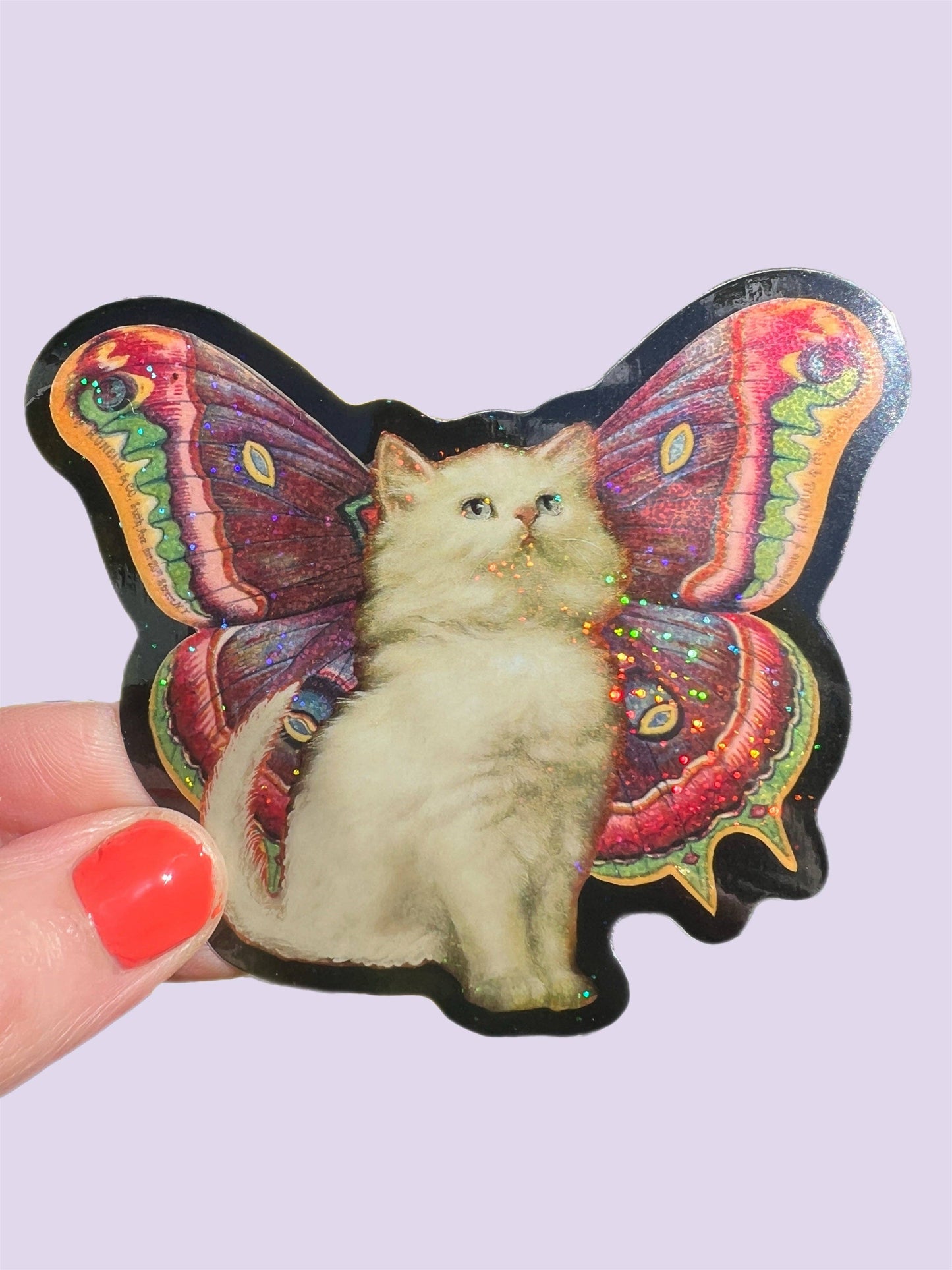 Sticker of a cat with butterfly wings held by a hand with red nail polish on a light purple background.
