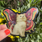Butterfly-shaped sticker with a cat design held in front of green plants and yellow flowers.