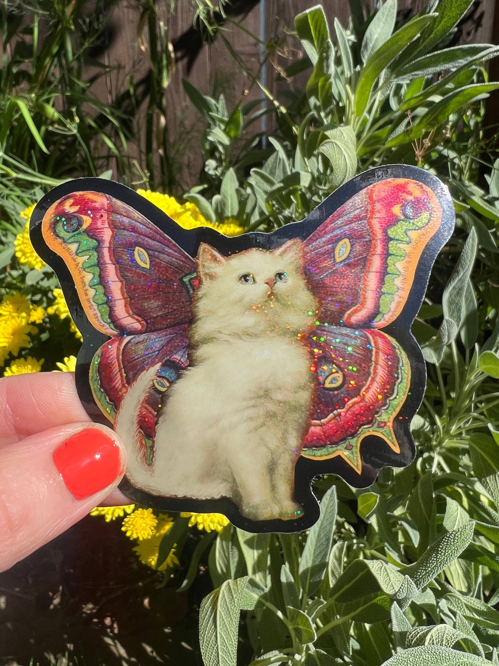 Butterfly-shaped sticker with a cat design held in front of green plants and yellow flowers.