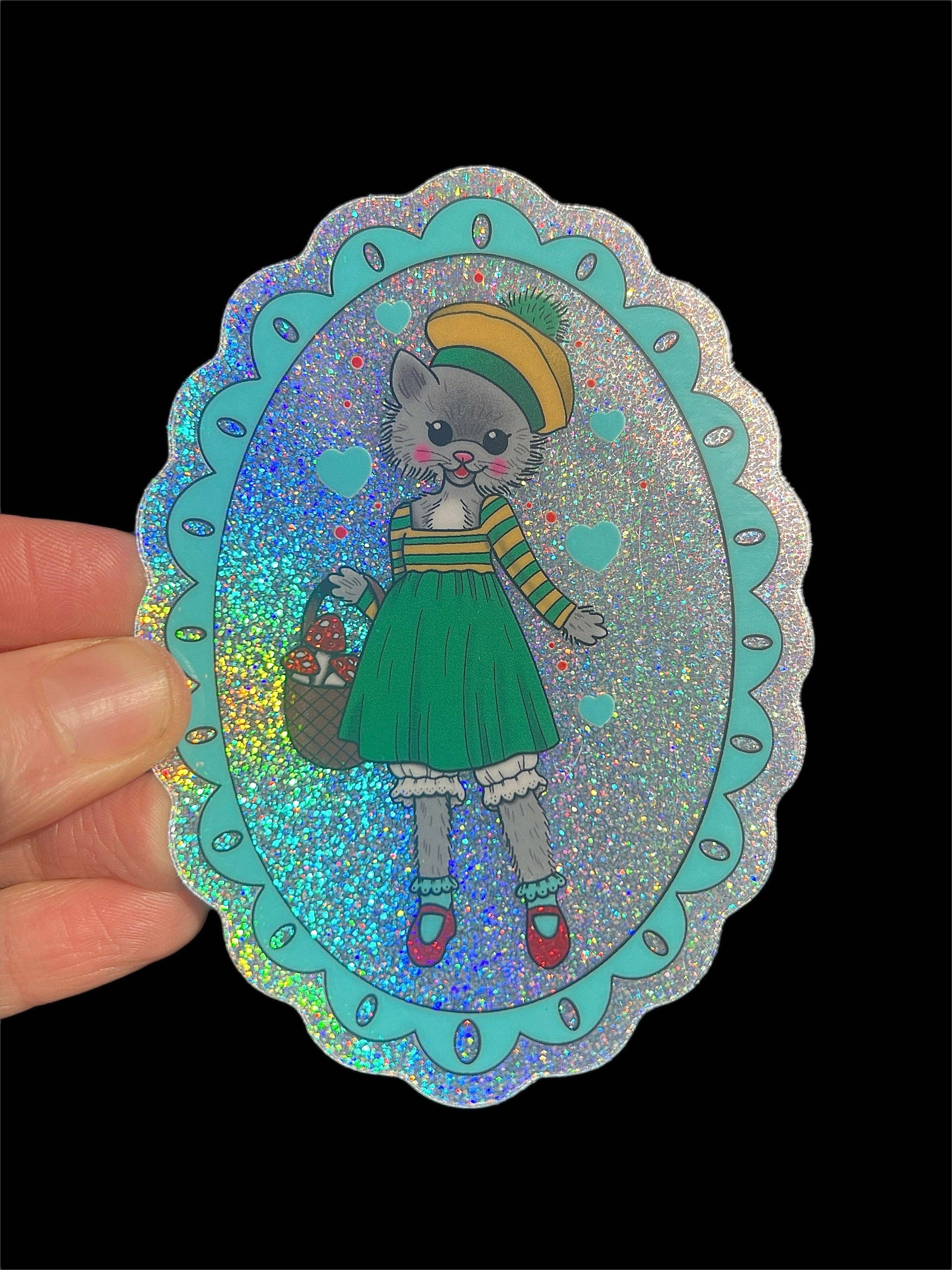 Sticker of a cat in a green dress with a colorful hat held against a black background