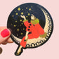 Decorative sticker with a person in a red coat and gold accents sitting on a crescent moon, surrounded by stars, held against a pink background.