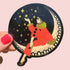 Decorative sticker with a person in a red coat and gold accents sitting on a crescent moon, surrounded by stars, held against a pink background.