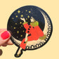 Decorative sticker of a person on a crescent moon with stars, held against a beige background.