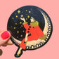 Sticker of a person in a red coat sitting on a crescent moon with stars, held against a pink background.