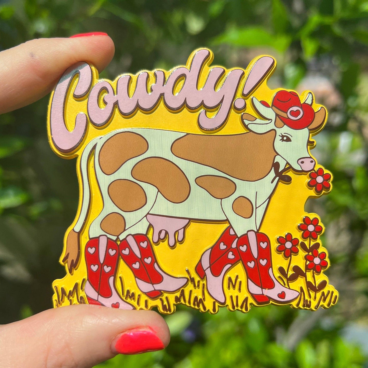 Colorful cow magnet with 'Cowdy!' text held by a hand against a green leafy background