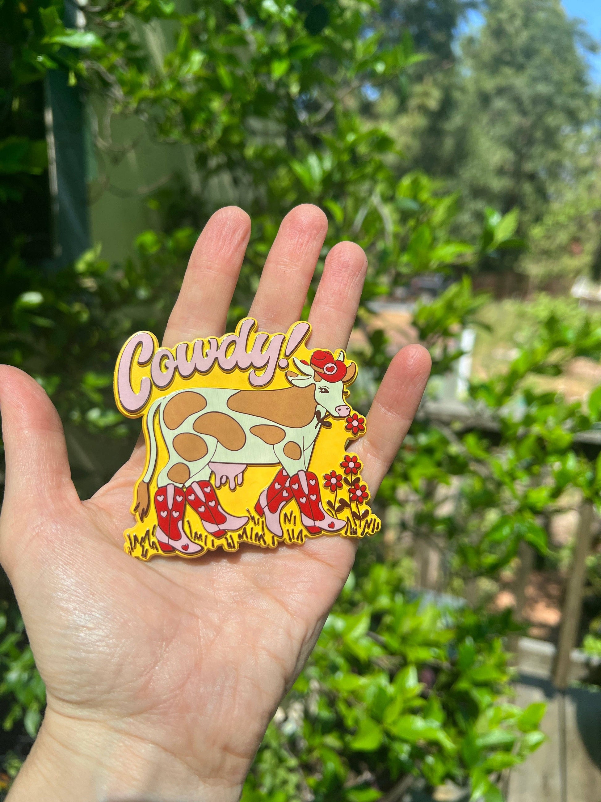 Hand holding a colorful cow magnet  with a blurred green outdoor background
