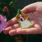 A hand holding a small illustrated patch or badge shaped like a dog wearing a party hat, with a green glittery outline, photographed outdoors against dark green foliage and a pink flower.