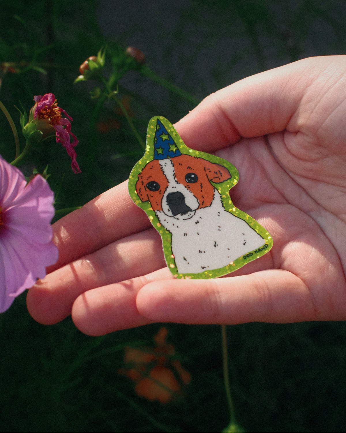 A hand holding a small illustrated patch or badge shaped like a dog wearing a party hat, with a green glittery outline, photographed outdoors against dark green foliage and a pink flower.