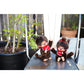 Two Monchhichi  with red bows on a metal surface with plants in the background.