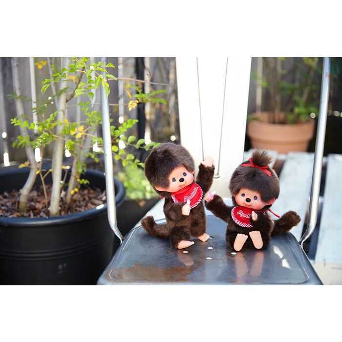 Two Monchhichi  with red bows on a metal surface with plants in the background.