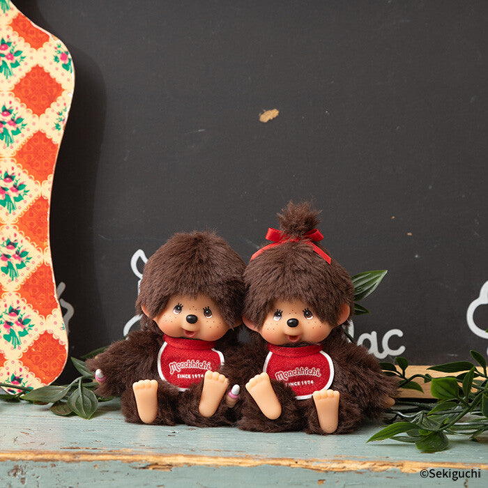 Two brown Monchhichi with red bows and shirts on a wooden surface with a dark background.