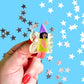 Hand holding a small fairy hair clip with a colorful hat against a blue background with star patterns.