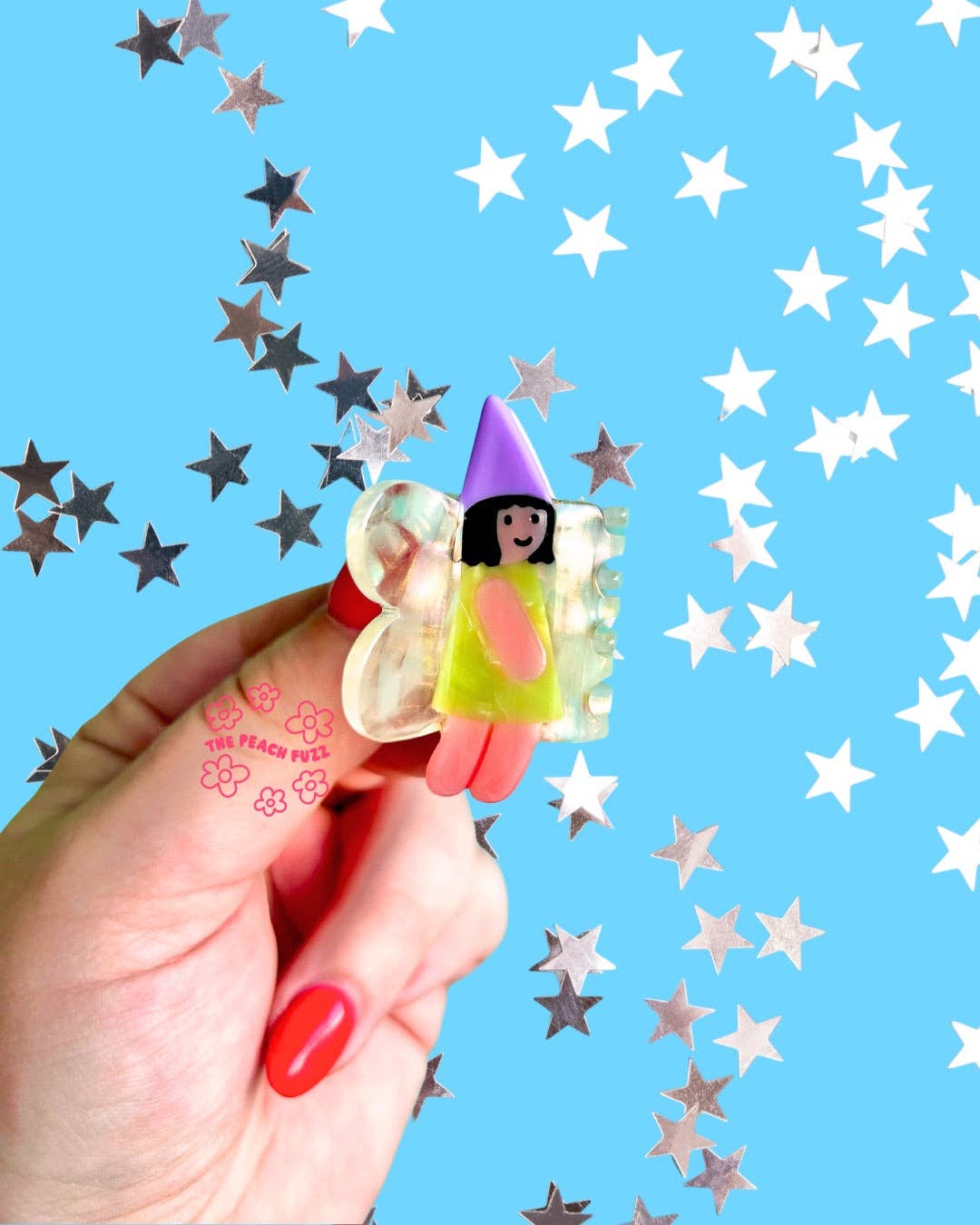 Hand holding a small fairy hair clip with a colorful hat against a blue background with star patterns.