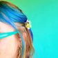 Person with blue hair and a decorative hair clip against a teal background
