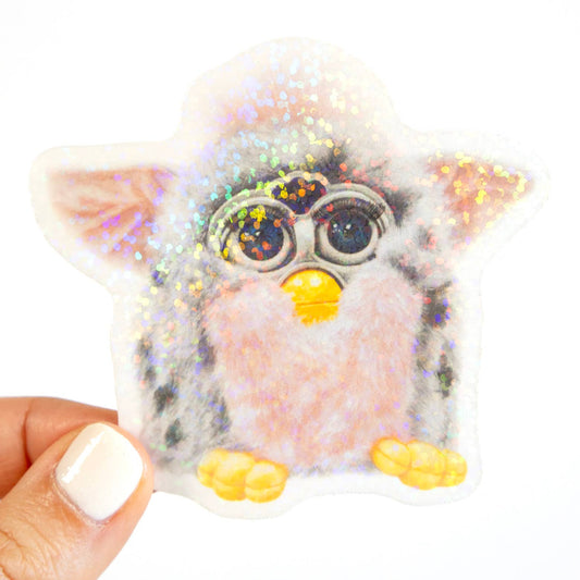 Glittery toy resembling a creature with large eyes and yellow feet, held by a hand.