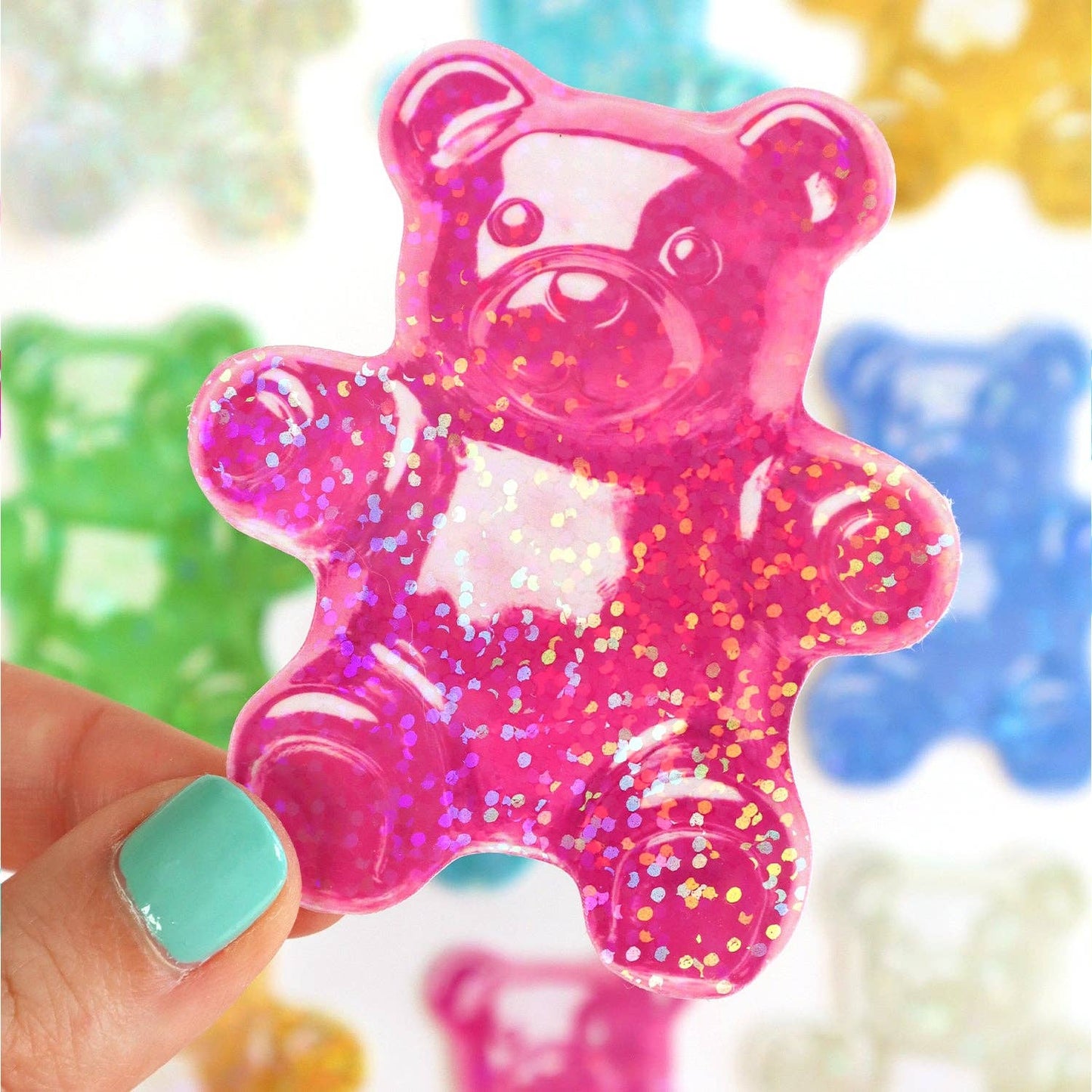 Pink glittery gummy bear sticker held by a hand with teal nail polish, colorful background.