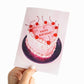 Hand holding a birthday card with a heart-shaped cake design and 'Happy Birthday!' text.