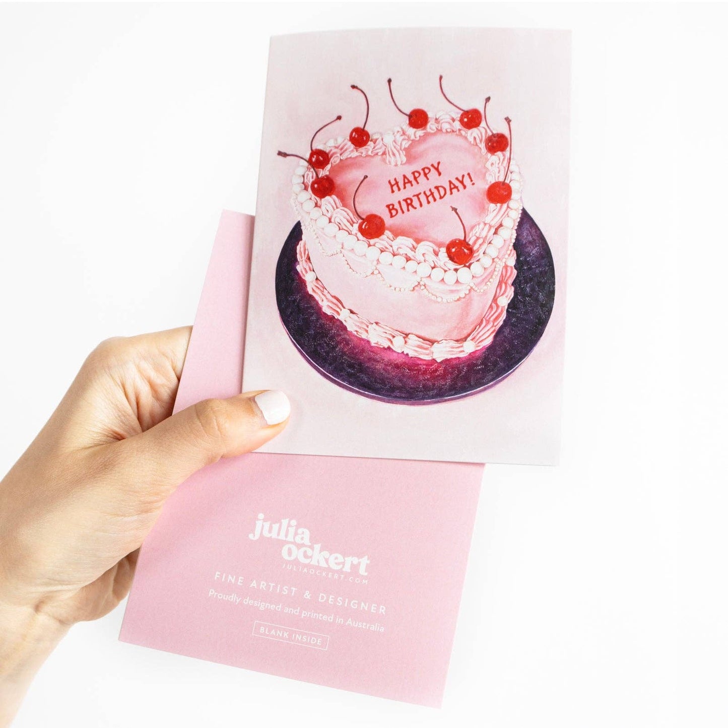 Hand holding a card with a birthday cake illustration and 'Happy Birthday!' text, brand 'Julia Ockert' visible.