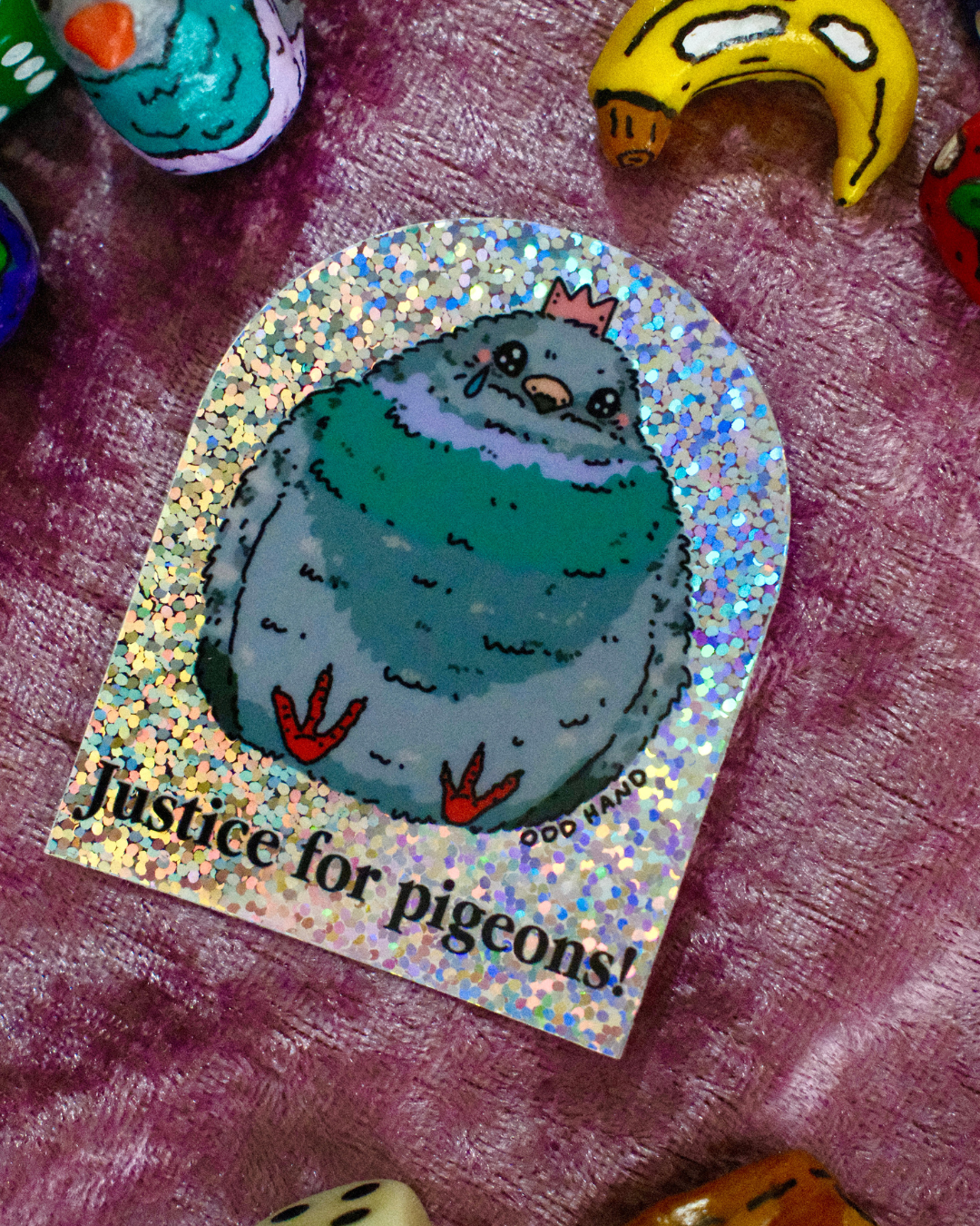A sticker featuring a cartoon pigeon with the text 'Justice for pigeons!' on a holographic background, placed on a purple textured surface with other colorful objects partially visible around the edges.
