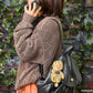 Person wearing a textured brown sweater with a black bag and teddy bear toy against a brick wall.