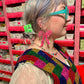 Person wearing colorful glasses and a beaded vest in front of pink shelves.