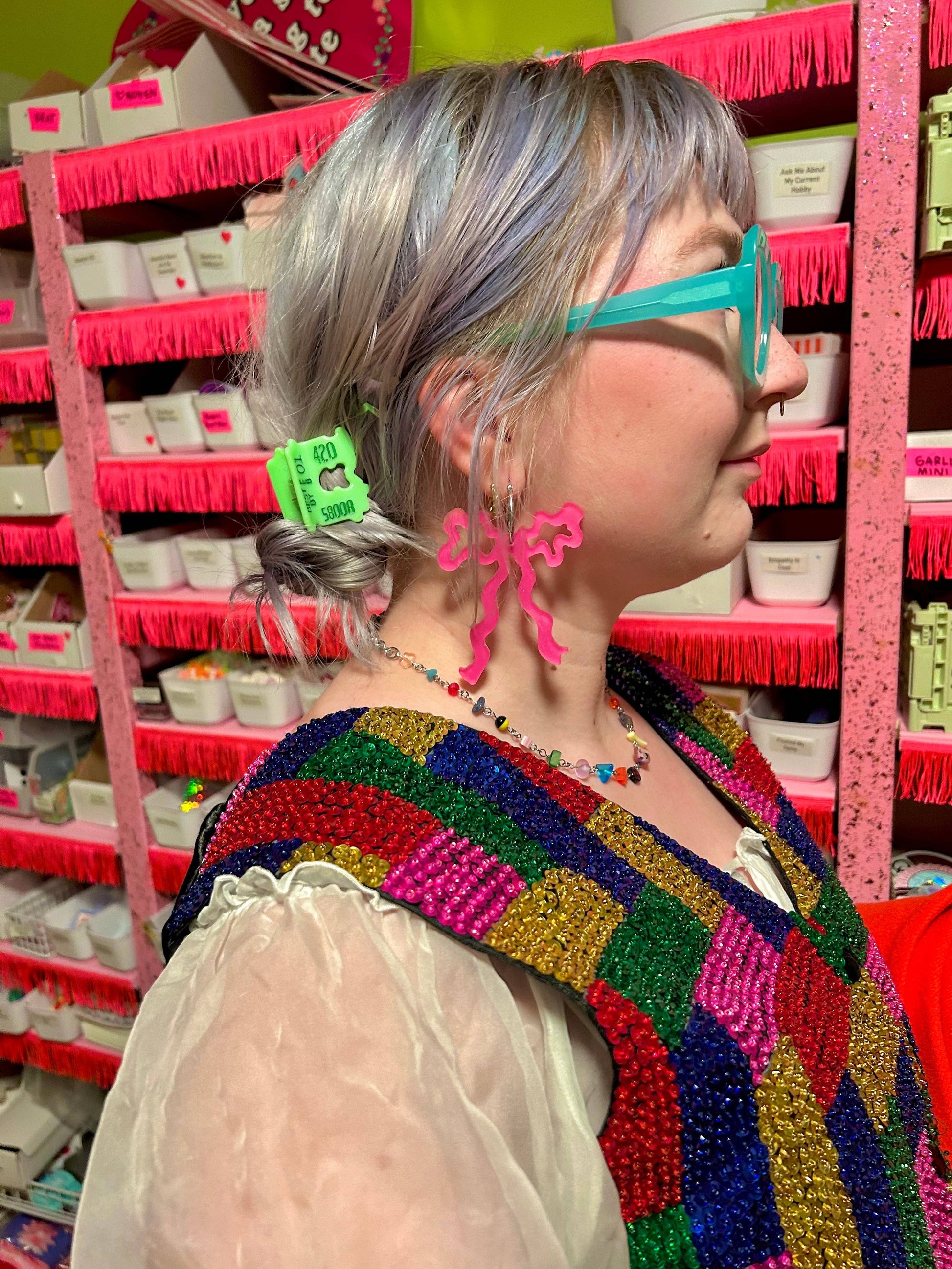 Person wearing colorful glasses and a beaded vest in front of pink shelves.