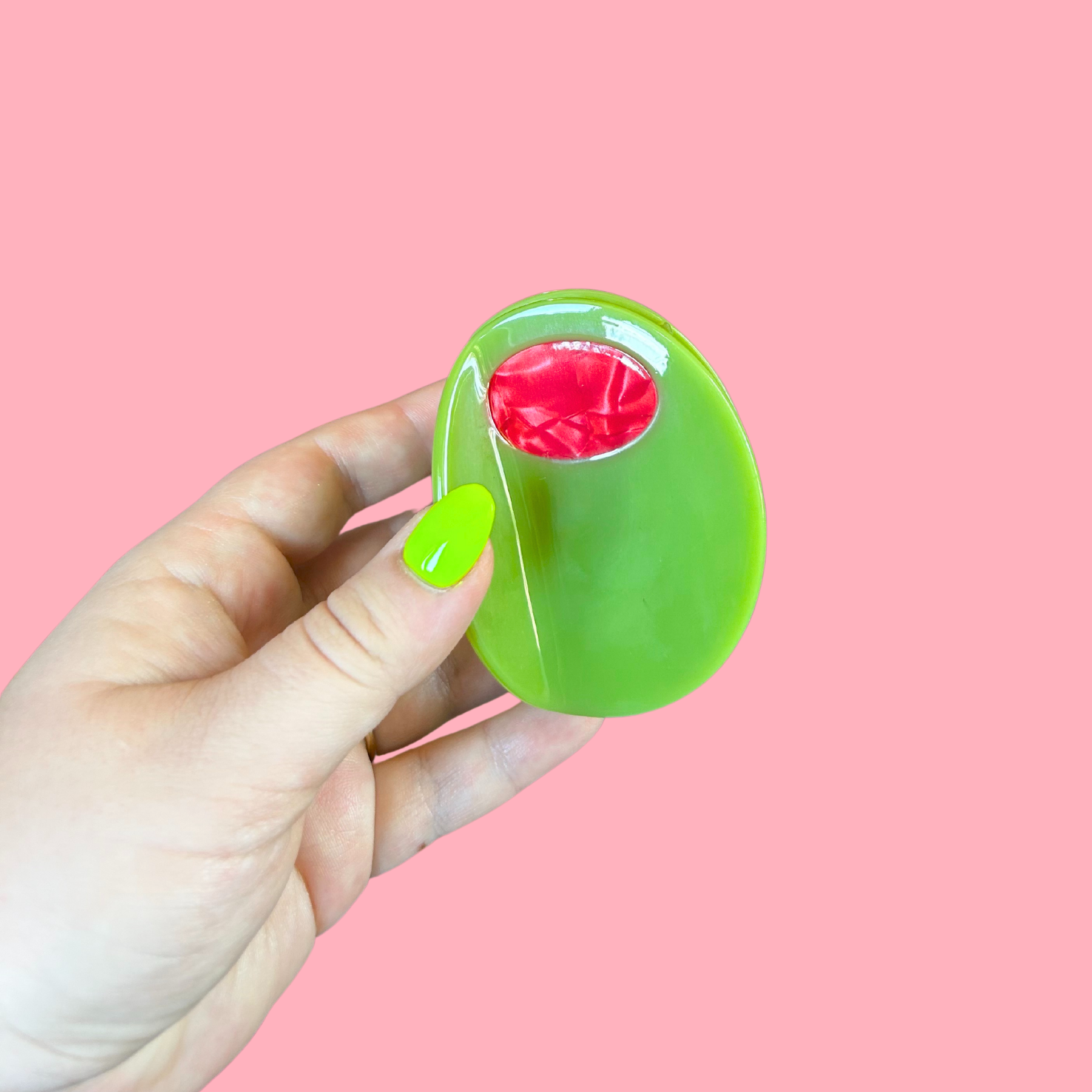 Hand holding a olive hair clip with a red center against a pink background