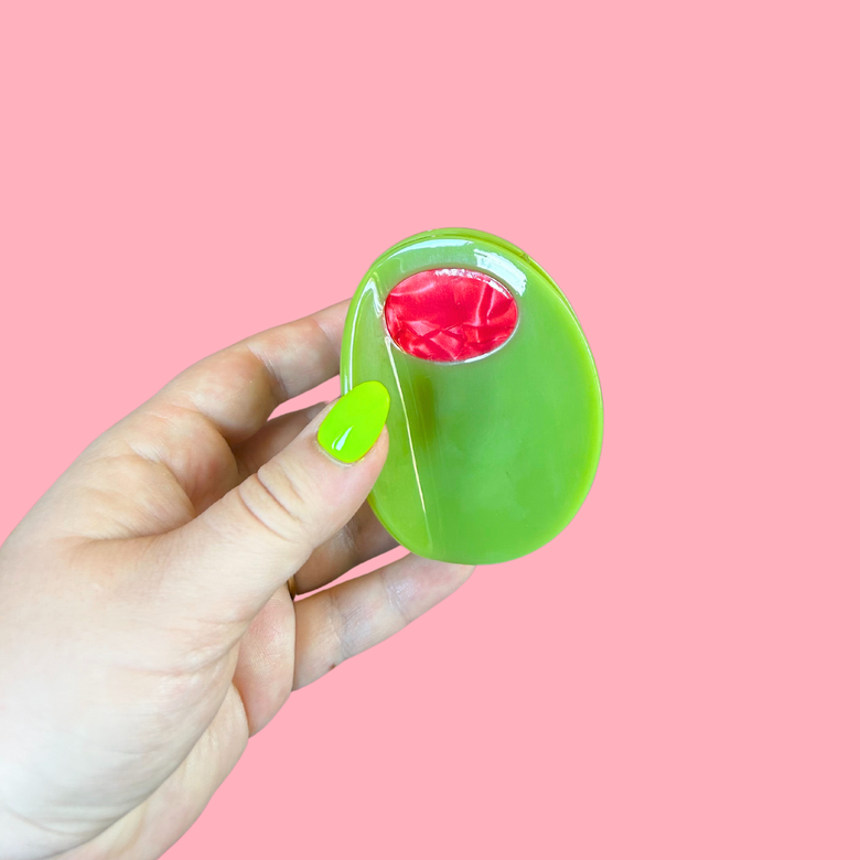 Hand holding a olive hair clip with a red center against a pink background