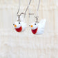 A pair of white bird-shaped dangle earrings with red/orange bibs, black eyes, orange beaks, and silver fishhook wires, shown against a soft beige textured background.