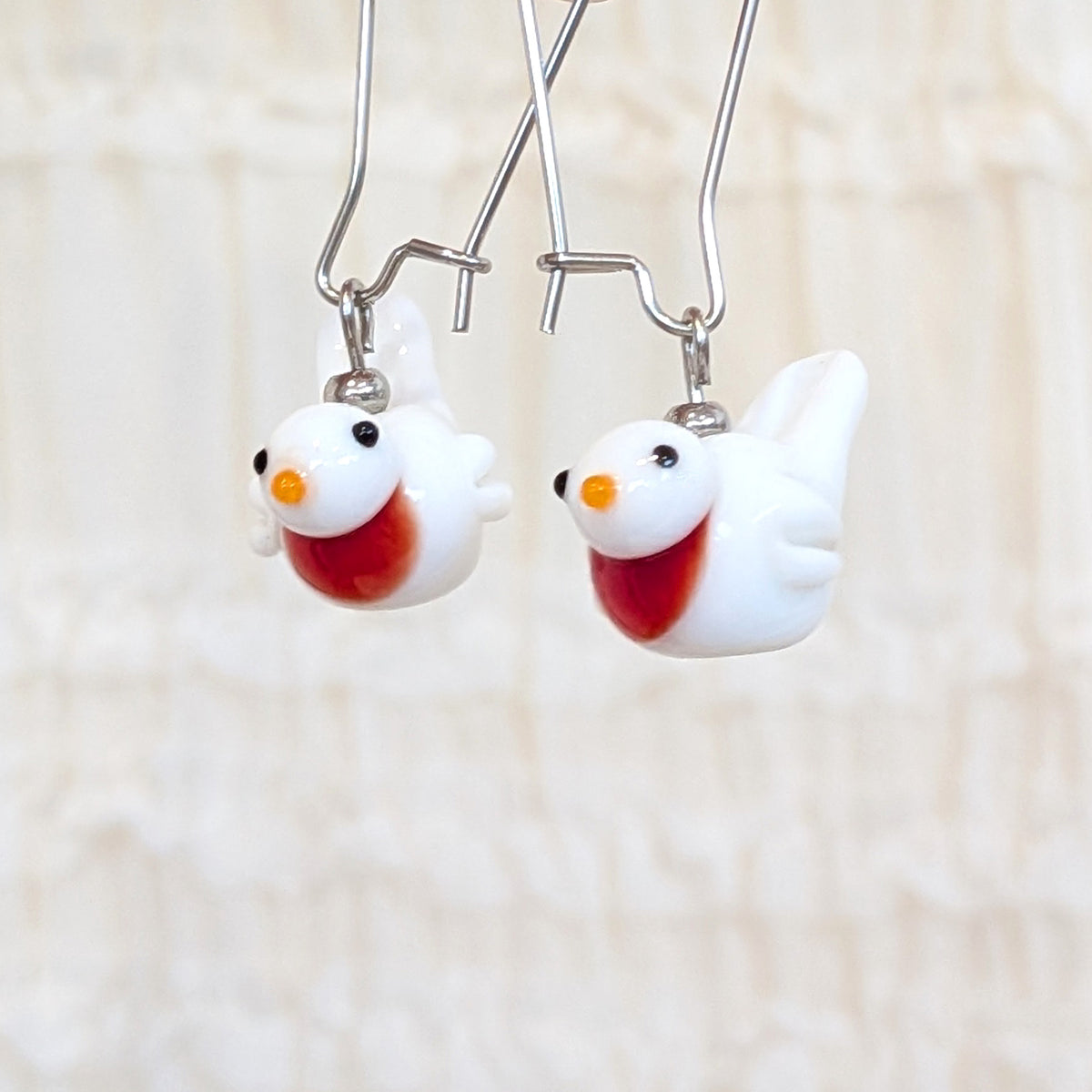 A pair of white bird-shaped dangle earrings with red/orange bibs, black eyes, orange beaks, and silver fishhook wires, shown against a soft beige textured background.