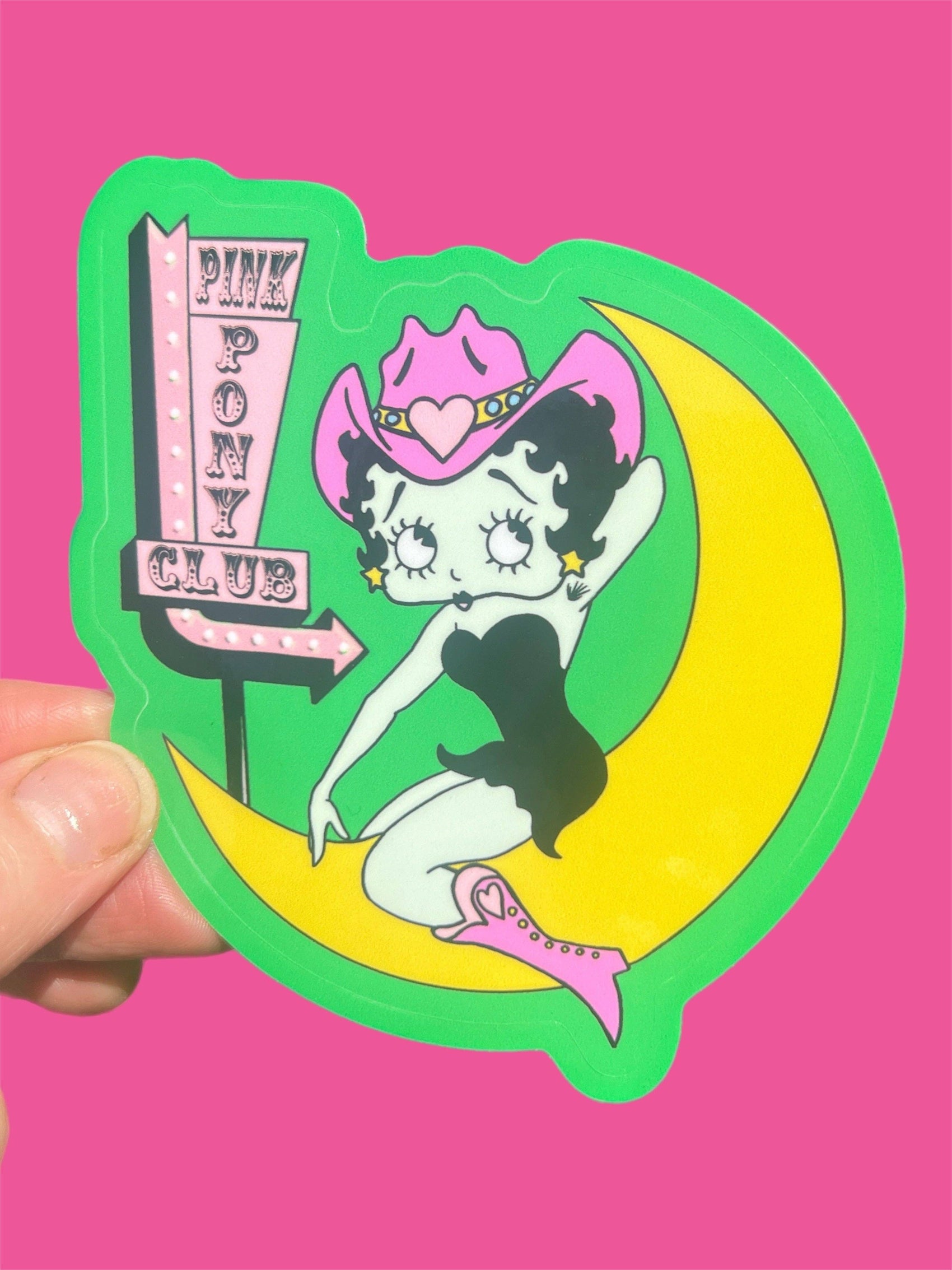 Sticker of betty boop on a moon with 'Pink Pony Club' sign, held against a pink background.